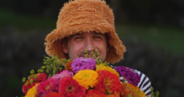 緑の庭の背景に赤 オレンジ 黄色の菊の花の花束を持っているパナマの帽子の白人男性は それを嗅ぎ 大きな目を作ります 高品質4Kビデオ映像 — ストック動画