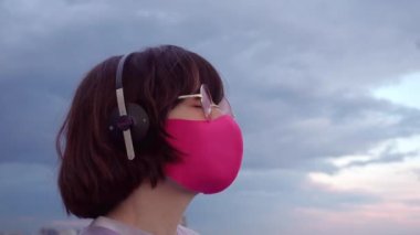 Pembe maskeli, güneş gözlüklü, kulaklıklı, dans eden, bulutlu gökyüzünde müzik dinleyen genç esmer kadın. Yüksek kalite 4k video kaydı