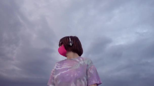 ピンクのマスクとヘッドフォンのダンスで若いブルネットの女性は 曇った空の背景に音楽を聞いている 高品質4K映像 — ストック動画