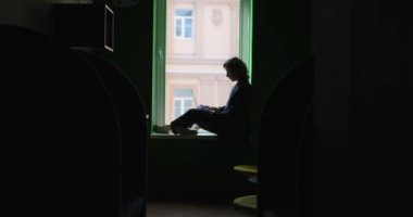 İş kadını çalışma konsepti. Çalışma sırasında açık ofis alanında pencere eşiğinde oturan, iş elbisesi giymiş, dizüstü bilgisayara bakan beyaz kadın silueti. Yüksek kalite 4k video