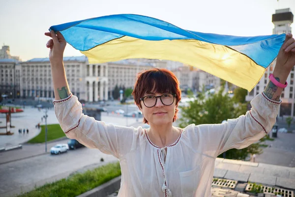 stock image Portrait of caucasian female in eyeglasses and white blouse with flag of Ukraine waving on wind with urban view on background. Patriotic woman with flag of Ukraine. Freedom concept. High quality photo