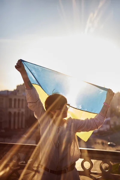 Caucasian female silhouette in white blouse with flag of Ukraine waving on wind with urban view on background and lens flares effect. Portrait of a patriotic woman with flag of Ukraine. Freedom time