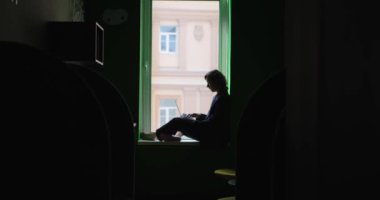 İş kadını çalışma konsepti. Çalışma sırasında açık ofis alanında pencere eşiğinde oturan, iş elbisesi giymiş, dizüstü bilgisayara bakan beyaz kadın silueti. Yüksek kalite 4k video