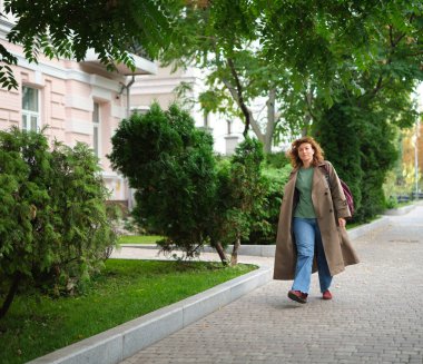 Genç, modaya uygun, modaya uygun, güzel, sıradan bir kadın sırt çantasıyla şehir caddesinde yürüyor. Kadın portresi, buluşmak için acele eden kot pantolon ve trençkot giyiyor. Yüksek kalite fotoğraf