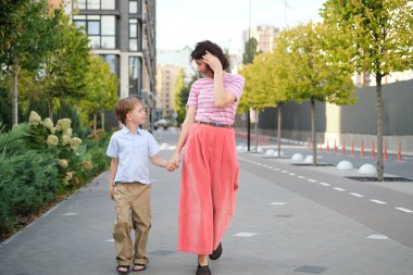 Beyaz, genç, güzel, mutlu bir anne ve sokakta yürüyen ve birbirine bakan sevimli küçük bir oğul. Kadın ve küçük çocuk gülümsüyor, konuşuyor ve tatil yapıyorlar. Yüksek kalite fotoğraf