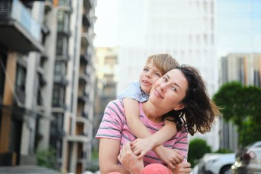 Modern mesken kompleksinde kameraya gülümseyen genç anne ve oğul. Birlikte eğlenmek, mutlu ebeveynlik, yeni ev konsepti. Şehir arka planında parlak bir yaz günü. Yüksek kalite fotoğraf