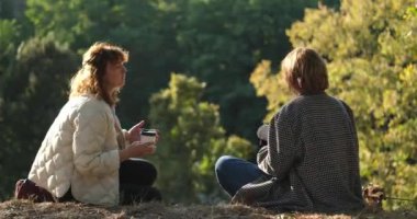 Parkta çimlerin üzerinde oturup gülen iki arkadaş. Süveter giymiş küçük bir köpekle paket kahve içmek. Güneşli akşam ışığı, hafta sonu konsepti. Yüksek kaliteli video görüntüsü 4k