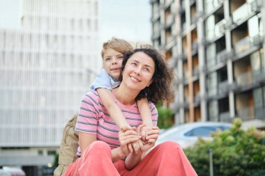 Modern mesken kompleksinde kameraya gülümseyen genç anne ve oğul. Birlikte eğlenmek, mutlu ebeveynlik, yeni ev konsepti. Şehir arka planında parlak bir yaz günü. Yüksek kalite fotoğraf
