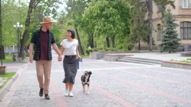 Barınak köpeği olan mutlu çift yazın eski şehrin arka planında sarılıyor. Siyah saçlı kadınlar ve şapkalı bir adam kurtarılmış köpeği olan sevimli, genç ve modern bir çift. Yüksek kaliteli video görüntüsü 4k