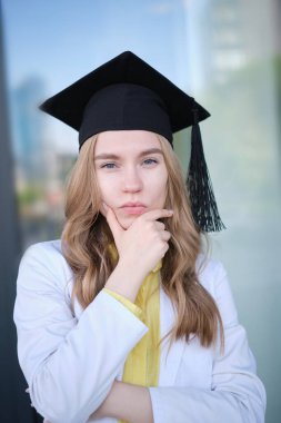 Genç, sarışın, tıp üniformalı, mezuniyet şapkalı, neşeli ve keyifli bir kadın. Dışarıda dikilmiş, sağlık ve veterinerlik eğitimimi bitirdiğim için mutluyum. Yüksek kalite fotoğraf