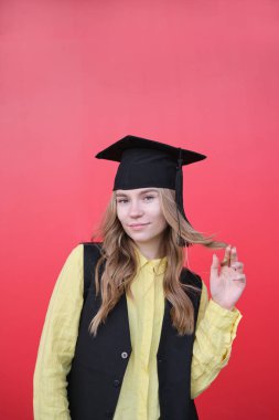 Mezuniyet şapkalı güzel sarışın kadın. Kırmızı arka planda duygusal bir yüz ifadesi var. Okulu, üniversiteyi ya da yüksekokul kalitesinde fotoğrafı bitirmekten mutlu.