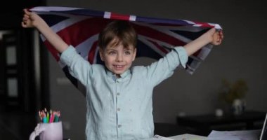 Mutlu çocuk gülümsüyor ve elinde İngiliz bayrağı, elinde dizüstü bilgisayar ve notlarla zt masasında oturuyor. Evde İngilizce öğrenme kavramı. video görüntüleri 4k