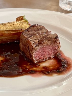 Orta pişmiş bir biftek, saf beyaz bir tabakta dinleniyor, üzerine kırmızı şarap sosu serpiştirilmiş..