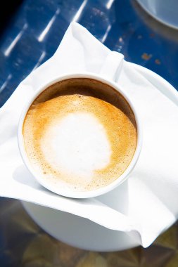 Parlak güneş ışığında sıcak bir fincan taze kapuçinonun üst görüntüsü. Sütlü beyaz peçeteli bir fincan sıcak kahve. Geleneksel İtalyan kahvaltı içeceği. Avrupa 'da tatil içkisi. Kapatın. Yüksek