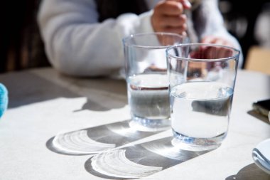 Bir insan, tahtadan bir masada rahatça oturuyor ve önünde iki bardak ferahlatıcı su duruyor.