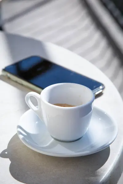 Buharı tüten bir fincan kahve modern bir masadaki zarif bir cep telefonunun yanında oturarak rahat bir atmosfer yaratıyor.