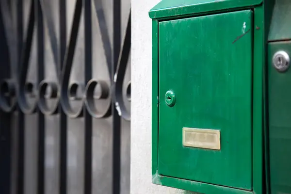 Canlı yeşil bir posta kutusu süslü dövmeli demir bir kapının yanına yerleştirilir ve bahçe alanında büyüleyici bir kontrast yaratır.