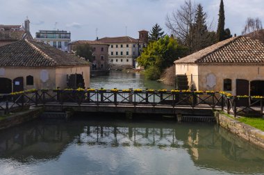 Portogruaro, İtalya (1 Mart 2025) - Portogruaro, Veneto, İtalya 'da Lemene nehri boyunca tarihi su değirmenleri. Döşeme çatılı iki taş bina dar su yolunu kuşatıyor. Sarı çiçeklerle süslenmiş ahşap bir köprü nehri geçer.