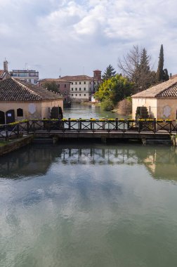 Portogruaro, İtalya (1 Mart 2025) - Portogruaro, Veneto, İtalya 'da Lemene nehri boyunca tarihi su değirmenleri. Döşeme çatılı iki taş bina dar su yolunu kuşatıyor. Sarı çiçeklerle süslenmiş ahşap bir köprü nehri geçer.