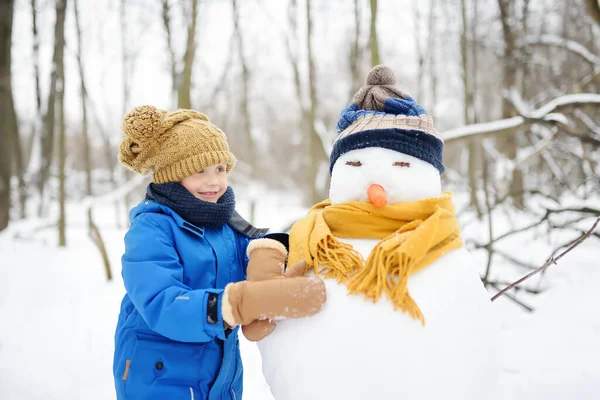 小男孩在雪地公园里堆雪人 孩子们抱着戴着帽子和围巾的雪人 冬天与有孩子的家庭一起积极地进行户外活动 孩子们在雪地的冬日公园里散步 — 图库照片