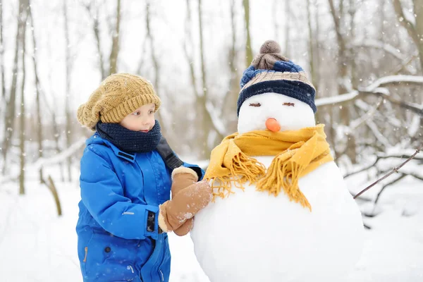 雪の公園に雪だるまを作る小さな男の子 帽子とスカーフを身に着けている雪だるまを受け入れる子供 冬に子供連れの家族とのアクティブな屋外レジャー 雪の冬の公園を散歩中の子供 — ストック写真