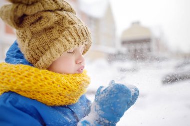 Küçük çocuk ellerinden kar üflüyor. Çocuk karlı bir günde parkta yürümekten zevk alır. Bebek kar yağarken eğleniyor. Çocuklu aileler için açık hava kış aktiviteleri.