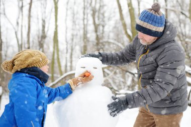Babasıyla küçük bir çocuk kardan adam yapıyor. Kışın çocuklu aileyle açık hava eğlencesi. Karlı bir kış parkında gezinirken ebeveyn ve çocuk