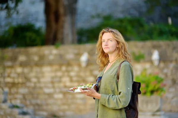 Portrait of charming young tourist woman in medieval town. Attractive red-haired and green eyes girl eating pizza. National european cuisine. Street food. Fast food or junk food to go.