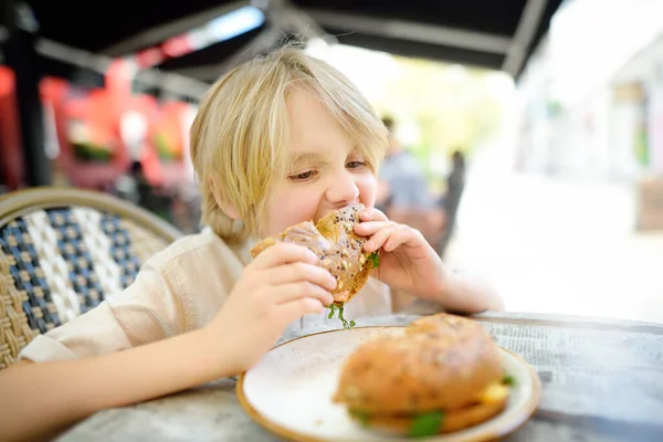 かわいいブロンドの男の子は 屋外のファーストフードレストランでサーモンとアルグラと大きなベーグルを食べています 子供のための健康的な食事 — ストック写真