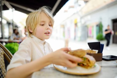 Açık hava fast food restoranında somon ve roka ile büyük simit yiyen sevimli sarışın çocuk. Çocuklar için sağlıklı yemek.