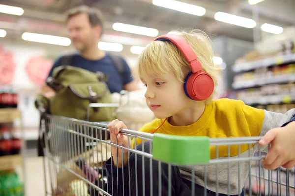 可爱的学龄前男孩带着耳机和播放器坐在食品店或超市的推车上 一个孩子正在听音乐或有声读物 而他的父母在购买食品杂货 和孩子一起购物 — 图库照片