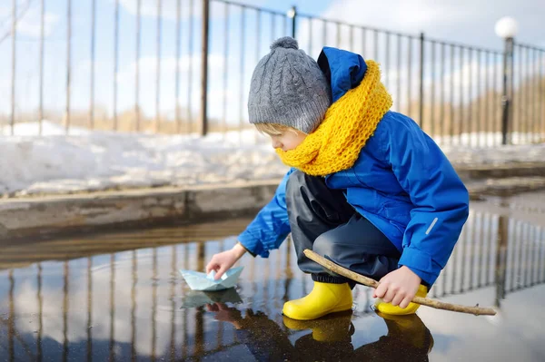 Little boy playing with origami paper boat in large puddle near town houses. Paper ship floating in water. Outdoor activities for kids in early spring