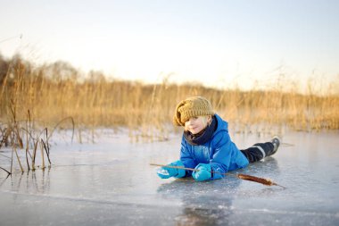 Tatlı anaokulu çocuğu soğuk, güneşli kış günbatımında donmuş bir gölün buzunda veya nehirde oynuyor. Çocuk aile yürüyüşü sırasında buz saçağı ve kuru sazlıkla eğleniyor. Çocuklar kışın açık hava oyunları