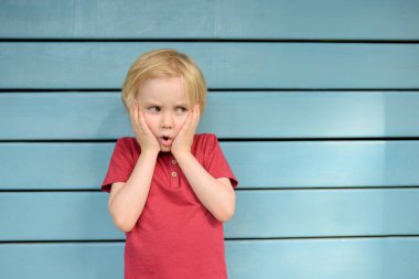 Kırmızı tişört giyen kızgın ve üzgün küçük çocuk mavi tahta arka planda duruyor. Yüz jesti.