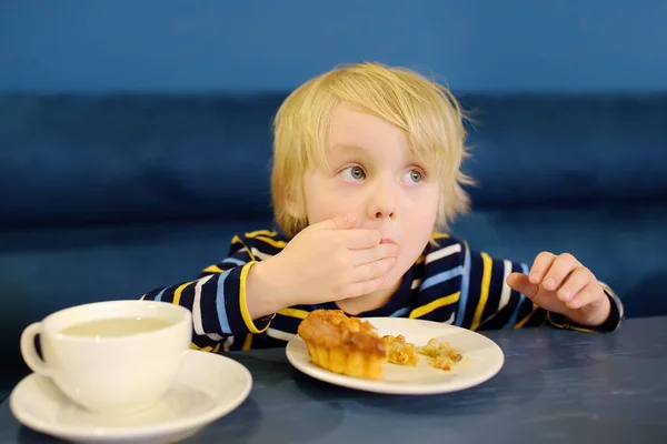 Cute blonde boy eating pie and drinking tea in cafe or restaurant. Child eating with his hands. Healthy meal for children.