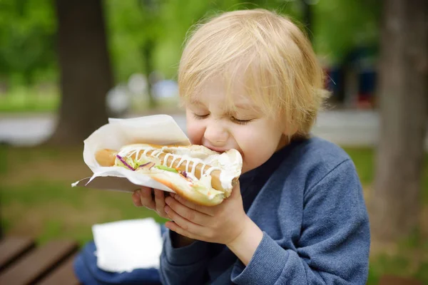 stock image Little boy eating hot dog in public park. Child enjoying his to go meal outside. Fast food is a junk food. Overweight problem kids.