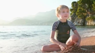 Deniz kıyısında oturan kumla ve taşlarla oynayan mutlu çocuk. Neşeli çocuk açık deniz kıyısında bir plaj tatili sırasında eğleniyor. Okul tatilinde çocuklar ve tüm aile için boş zaman.