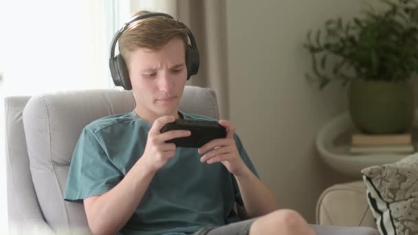 十几岁的男孩在玩一个便携式游戏机 一个青少年坐在客厅的椅子上玩小玩意 博彩青年 — 图库视频影像