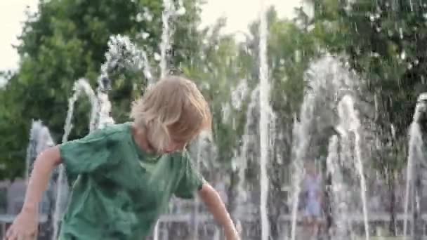 晴れた夏の日には噴水のウォータージェットの間の広場で小さな男の子が遊んでいます 大都市の子供たちのためのアクティブな夏のレジャー — ストック動画