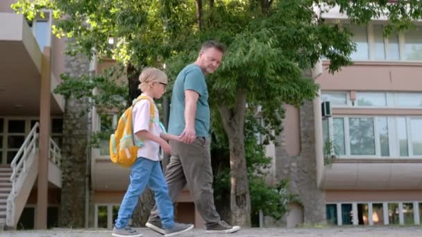 小学校の生徒が父親と一緒に学校を歩いている 少年は休日の翌日に学校の日がどうなったかを父親に話す 両親を持つ小学生 — ストック動画