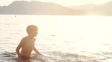 Mutlu çocuk gün batımında denizde yüzer. Neşeli çocuk gölde yüzerken su sıçrıyor ve eğleniyor. Okul tatilinde çocuklar için aktif boş zaman