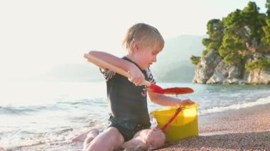 Deniz kıyısında oturan kumla ve taşlarla oynayan mutlu çocuk. Neşeli çocuk açık deniz kıyısında bir plaj tatili sırasında eğleniyor. Okul tatilinde çocuklar ve tüm aile için boş zaman.