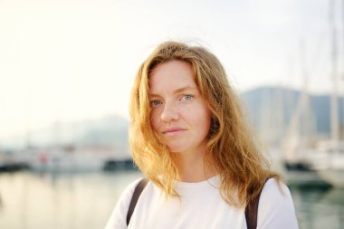 Portrait of charming young tourist woman on the Mediterranean coast. Attractive red-haired girl hiking by the sea shore. Tourism, adventure and travel concept.