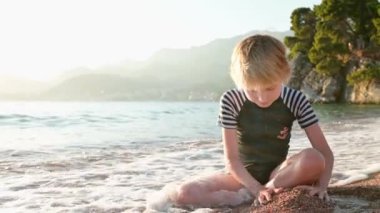 Deniz kıyısında oturan kumla ve taşlarla oynayan mutlu çocuk. Neşeli çocuk açık deniz kıyısında bir plaj tatili sırasında eğleniyor. Okul tatilinde çocuklar ve tüm aile için boş zaman.