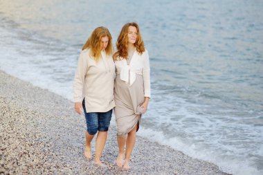 Kızıl saçlı kıdemli anne ve yetişkin hamile kızı deniz kıyısında birlikte yürüyorlar. Mutlu anne ve yetişkin çocuğu konuşuyor ve el ele tutuşuyor..