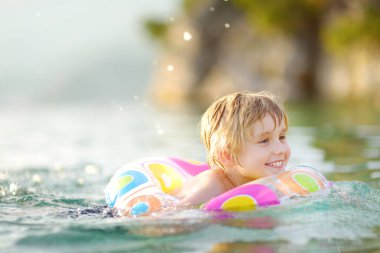 Küçük çocuk güneşli yaz gününde denizde yüzen renkli bir yüzükle yüzüyor. Temiz suda oynayan tatlı bir çocuk. Aile ve çocuklar yaz tatillerinde tatil yaparlar. Deniz kenarında dinlenme ve dinlenme.