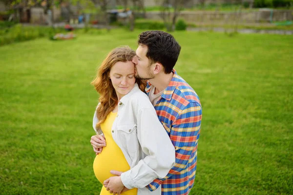 若い家族は赤ん坊を期待している 両親の幸せなカップル 一緒に歩くこと 妊娠中の女と抱き合ってキスするハンサムな男 夫は優しく妻を抱きかかえ 彼女の腹を楽しんで — ストック写真