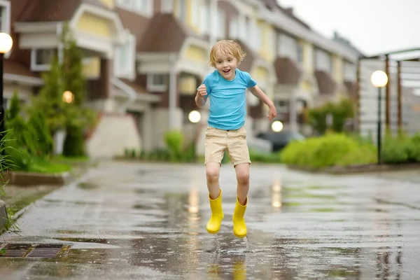 小さな町で雨の夏の日に水の水たまりにジャンプ黄色のゴムブーツを着て小さな男の子 子供は楽しんでる 雨の中で子供のための屋外ゲーム — ストック写真