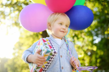 Sevimli küçük çocuk bir arkadaşını doğum gününde tebrik edecek. Bebek elinde renkli balonlar ve kutuda hediyeler tutuyordu. Anaokulu çocukları güneşli yaz parkında açık hava partisini kutluyor.