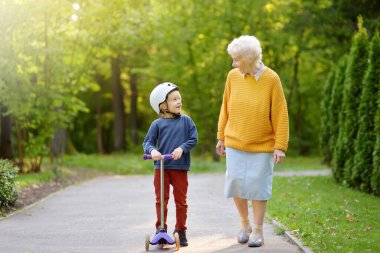 Güzel büyükanne ve küçük torunu sonbahar parkında birlikte yürüyorlar. Scooter kullanan bir çocuk. Aktif aile eğlencesi.